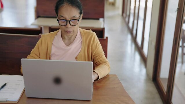 Asian senior woman types text working with laptop computer at home.