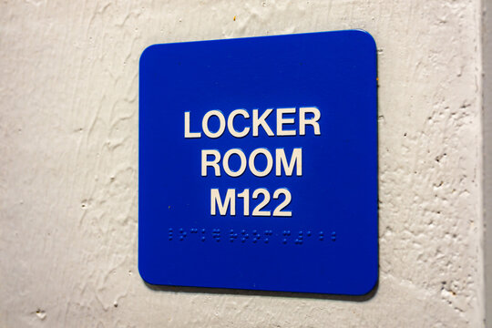 Blue LOCKER ROOM M122 sign with braille, on a white block wall.	