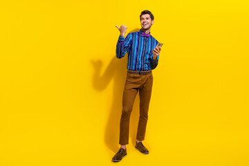 Full body photo of optimistic man wear bow tie hold smartphone look directing at offer empty space isolated on yellow color background