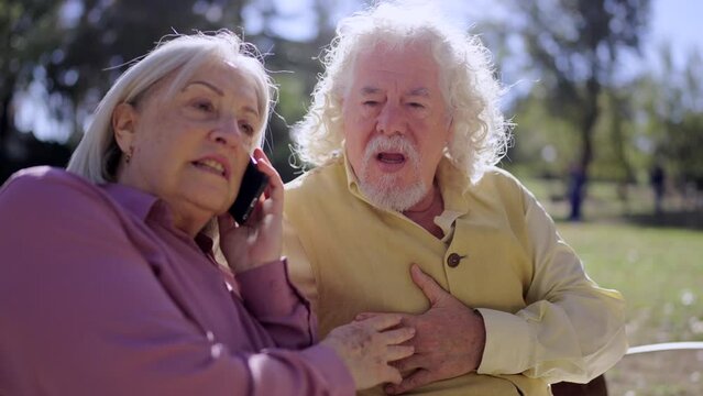 Video of a worried mature woman calling emergencies while senior man suffering a heart attack in a park