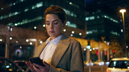 Woman scrolling smartphone night street. Successful late businesswoman wait taxi