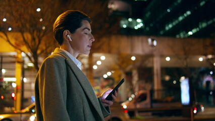 Woman walking street earphones at urban lights. Upset executive listen music