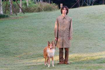 Portrait of a handsome Chinese young man in khaki overcoat with his rough collie dog on green grass...