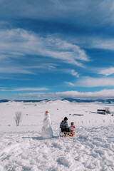 Fototapeta na wymiar Mom with a small child sit on a sled near a snowman and look at the snow plain. Back view