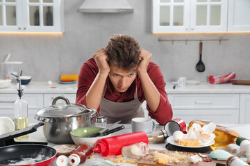 Upset man in messy kitchen. Many dirty dishware, utensils and food leftovers on table