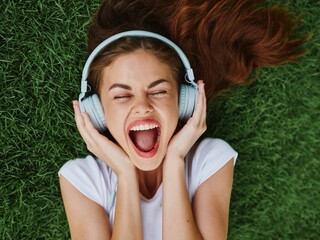 Beautiful woman in headphones listening to music lying on the grass green lawn in the park in the summer and singing with her mouth open, happiness in nature