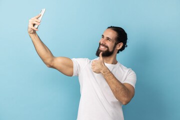 Portrait of man with beard wearing white T-shirt showing thumbs up like gesture and winking looking...