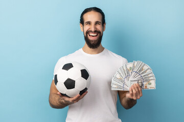 Portrait of man with beard wearing white T-shirt holding soccer ball and lots dollars banknotes,...