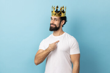 Portrait of proud man with beard wearing white T-shirt and golden crown and pointing himself, looking at camera with smile, superior privileged status. Indoor studio shot isolated on blue background. - obrazy, fototapety, plakaty