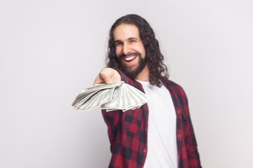 Portrait of cheerful optimistic bearded man with long curly hair in checkered red shirt giving to you big fan of money, you are winner. Indoor studio shot isolated on gray background.