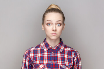 Portrait of surprised amazed shocked woman with bun hairstyle looking at camera with big eyes, sees something astonished, wearing checkered shirt. Indoor studio shot isolated on gray background.