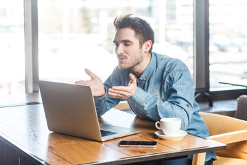 Portrait of positive romantic young man freelancer in blue jeans shirt working on laptop, having video call, sanding air kisses over palms. Indoor shot near big window, cafe background.