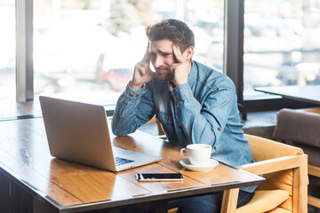 Portrait of tired exhausted sick man freelancer in blue jeans shirt working on laptop, spending long hours in front of computer screen, massaging temples. Indoor shot near big window, cafe background.