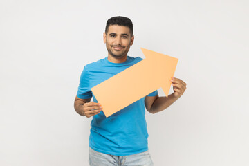 Smiling attractive unshaven man wearing blue T- shirt standing holding paper arrow indicating aside, looking at camera with happy expression. Indoor studio shot isolated on gray background.