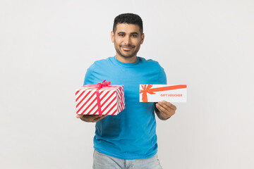 Portrait of pleased friendly unshaven man wearing blue T- shirt standing holding present box and gift voucher, looking smiling at camera. Indoor studio shot isolated on gray background.