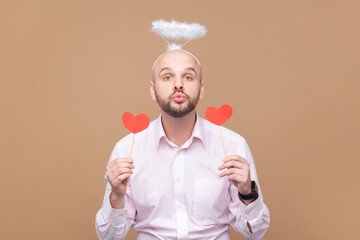 Portrait of flirting romantic bald bearded man with nimb over head holding red little hearts, sending air kisses, wearing light pink shirt. Indoor studio shot isolated on brown background.