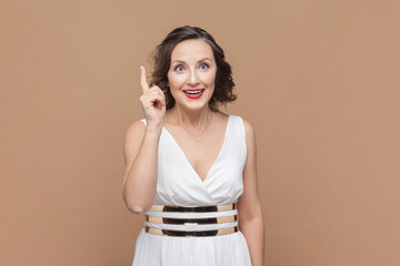 Portrait of inspired middle aged woman with wavy hair pointing finger up with genius idea, surprised by suddenly invented smart solution. Indoor studio shot isolated on light brown background.