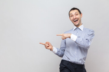 Portrait of extremely happy overjoyed handsome man pointing with both index fingers aside at copy space for advertisement, wearing light blue shirt. Indoor studio shot isolated on gray background.