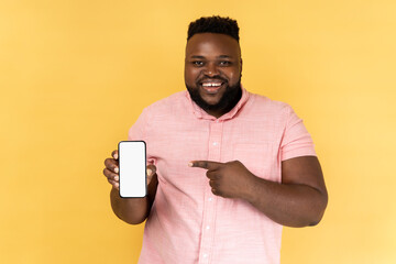 Portrait of cheerful man wearing pink shirt pointing at cell phone screen, looking at camera,...