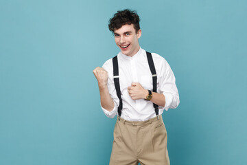 Portrait of extremely happy positive young handsome man wearing white shirt and suspender clenched...