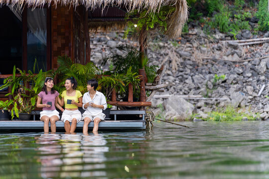 Group of Young Asian woman sitting on lake house balcony with splashing legs in water and talking together. People enjoy and fun outdoor lifestyle travel nature mountain on summer holiday vacation