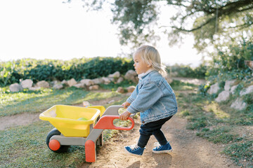Little girl with a toy wheelbarrow walks along a dirt path in the park - obrazy, fototapety, plakaty