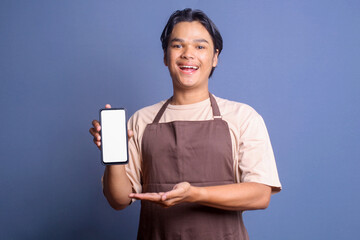 Young Asian man wear barista apron showing blank phone screen for mockup with happy expression
