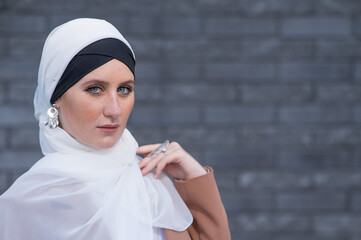 Portrait of a young blue-eyed woman in a hijab against a gray brick wall. 