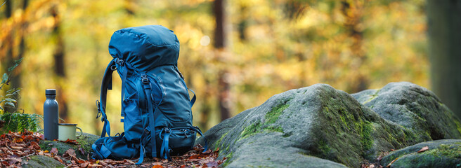 Hiking equipment in forest. Blue backpack, thermos and travel mug on rock. Panoramic view