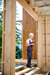 Carpenter constructing wooden framed house. Man worker cladding facade of house, using a screwdriver, wearing work overalls and helmet. Concept of modern eco-friendly construction.