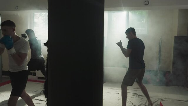 Fighters train hits on punching bags in gym. Professional sportsmen team does punches and kicks in smoky sports club slow motion. Athletic workout