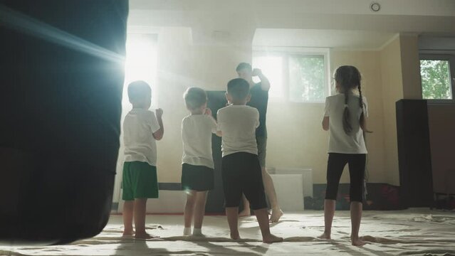 Trainer demonstrates punching to kids at martial arts school. Junior athletes repeat hits with hands following master in gym slow motion. Healthy activity