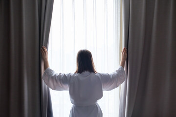Young woman in white bathrobe stands at the window and opens the curtains with her hands close-up, rear view.