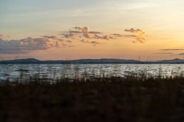 Morning waterfront view of Sirindhorn Dam Beach. We can see beach grass, river waves, and mountains.