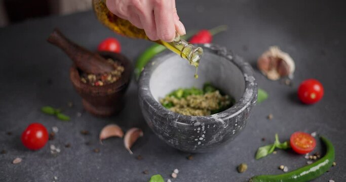 Woman pours cooking olive oil into stone mortar with green sauce chopped ingredients