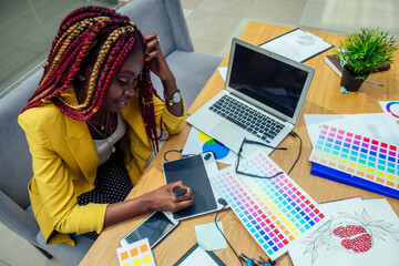 woman fashion designer with afro pigtails dreadlocks working on her atelier or print publishing house and graphic designer draws a sketch on a tablet. cosmetic distributor working in the office - obrazy, fototapety, plakaty