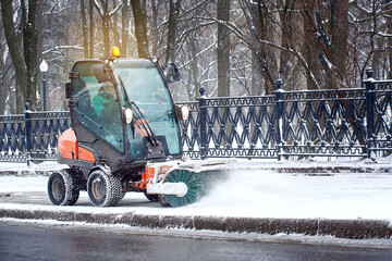 Small vehicle with rotating brush sweeping snow from sidewalk. Small tractor brushing snow from...