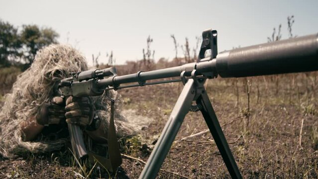 A close-up of a sniper wearing a camouflage suit or grass cloak, waiting and looking through a rifle scope. The sniper aims and shoots. Marksmanship shooting. Cinematic shot
