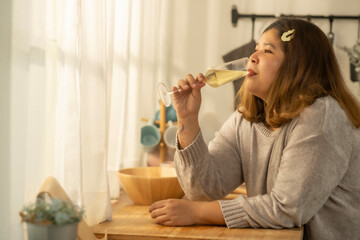 Young beautiful smiling over weight woman sitting in kitchen with glass of white wine on a sunny day - food, beauty, spring concept.