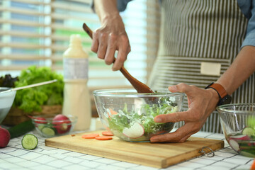 Cropped shot of middle age man in apron mixing fresh organic vegetables in glass bowl. Healthy...