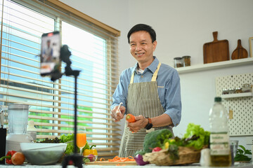 Cheerful middle age man in apron recording vlog on smartphone and sharing vegan cooking tips.