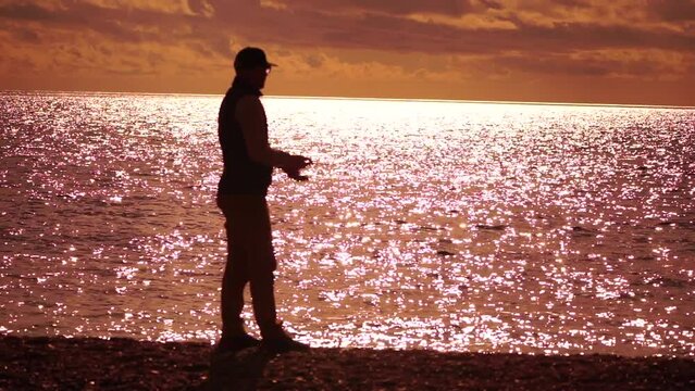 man standing on pier, holding a fishing rod and preparing to cast it into sea ocean at warm sunset. Man hobby fishing on sea tightens a fishing line reel of fish summer. Calm surface sea. Slow motion.