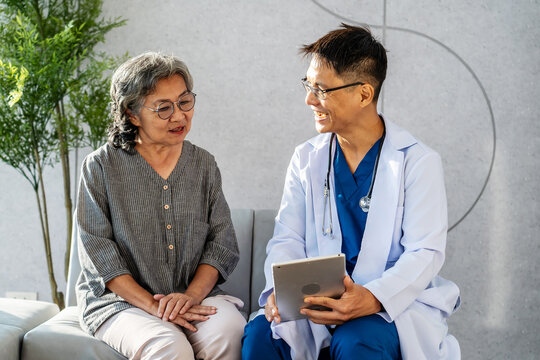 Asian Young Man Doctor Holding Digital Tablet Consulting Old Patient, Male Doctor Therapist In Uniform Sit On Sofa At Hospital Or Clinic With Senior Patient During Medical Visit Doing Health Checkup