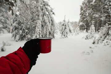 A hand holds a red coffee mug in a snowy winter forest