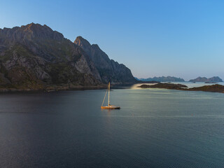 A sailboat under the midnight sun, behind a large hill in Lofoten, near Henningsvaer, in the...
