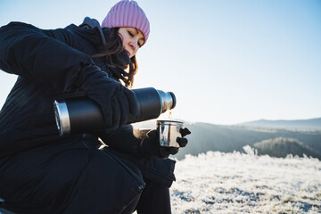 Girl pouring hot tea into a mug while sitting in winter in the forest against the background of mountains, winter trekking, warming drink, outdoor recreation, drinking morning coffee.