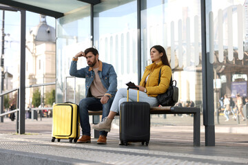 Being late. Worried couple with suitcases sitting at bus station outdoors - obrazy, fototapety, plakaty