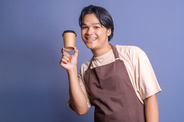 Young Asian barista guy in uniform showing takeaway coffee cup on blue background