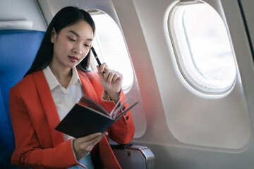 Smiling Asian woman enjoying reading a book comfortable flight while sitting in the airplane cabin, Passengers near the window.