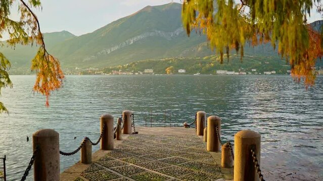 A pier near the water on Lake Como through the branches of yellowed larch against the backdrop of mountains at sunset